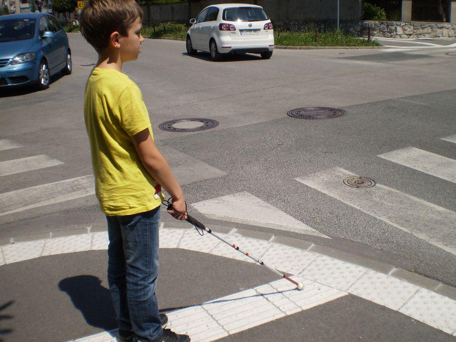 Na fotografiji na križišču dveh ulic stoji deček, ki ga vidimo od strani. Z belo palico tiplje po talnih taktilnih oznakah. Foto: Arhiv ZSSM