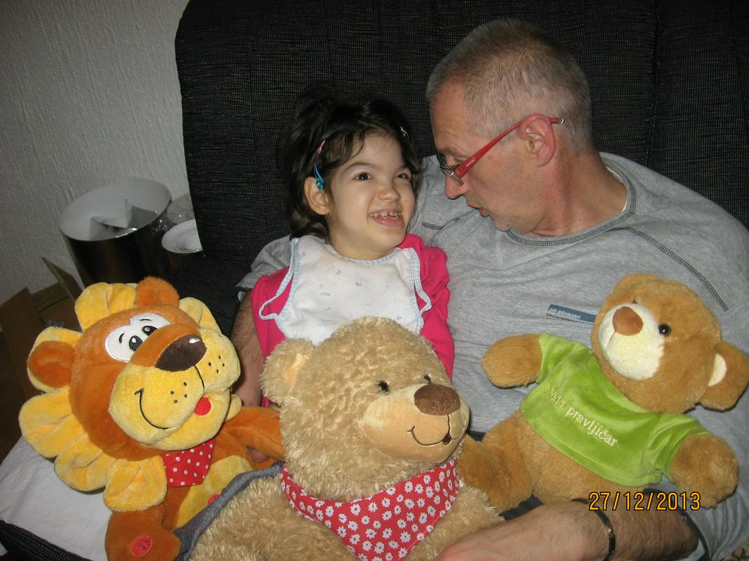 Opis fotografije: Na fotografiji deklica Lea sedi v očkovem naročju, obkrožena sta s plišastimi živalmi. Foto: osebni arhiv družine Murgel