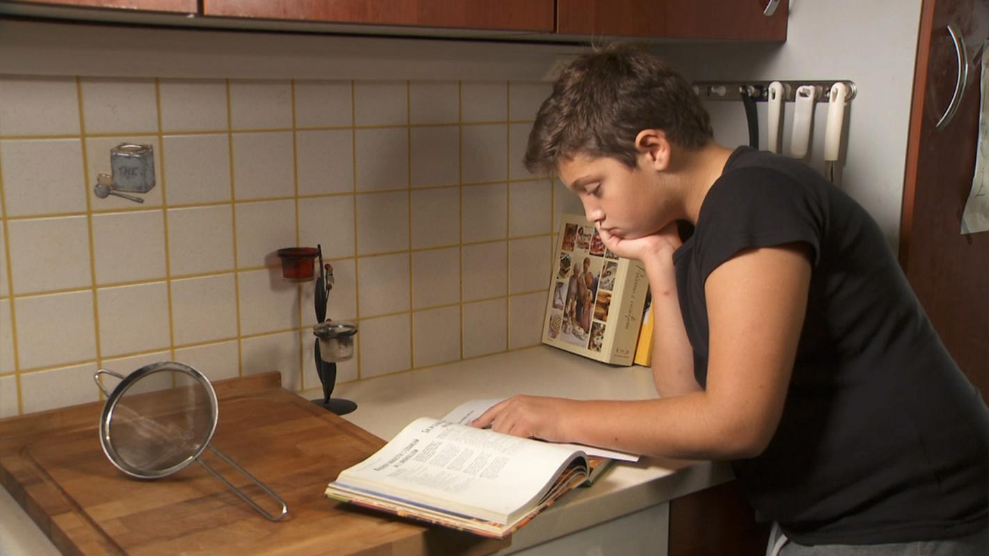 Na kuhinjski pult se naslanja deček in bere knjigo z recepti. Foto: arhiv oddaje Infodrom, Čhavoro, MMC RTV SLO