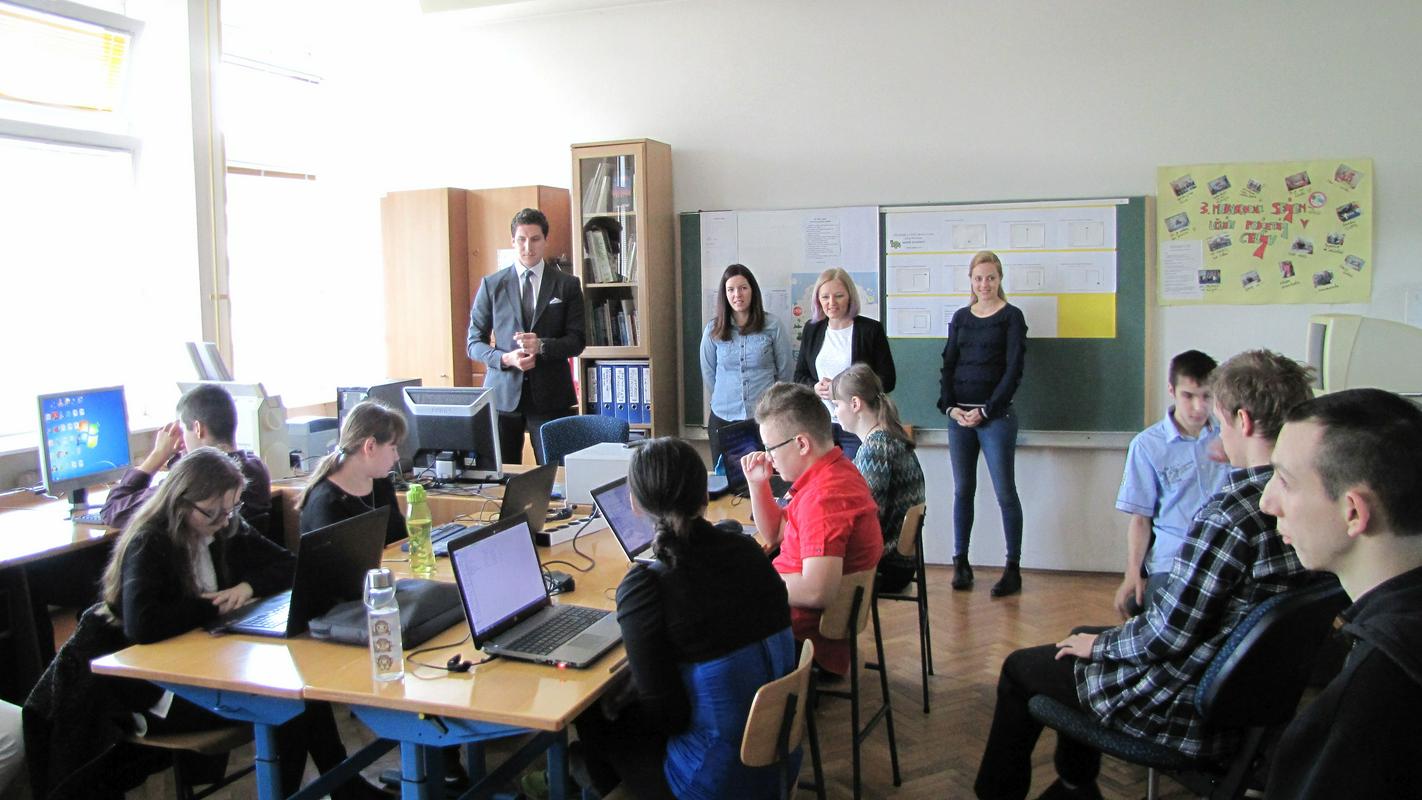 V učilnici pred računalniki sedijo dijaki. Pred tablo stojijo predavatelji. Fotografija: arhiv AmCham Slovenija.