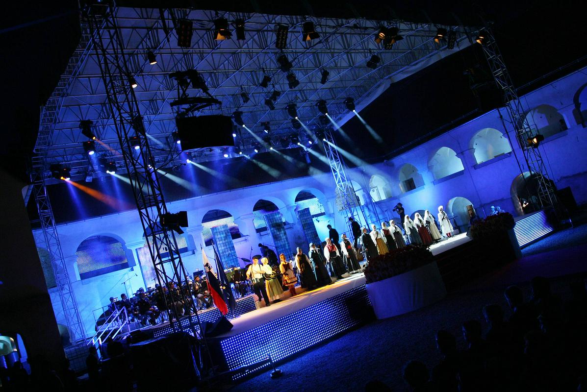 Nastopajoči v tradicionalnih prekmurskih oblačilih na pokritem odru pod sojem luči plešejo in pojejo. Foto: Andrej Križ/Ministrstvo za kulturo