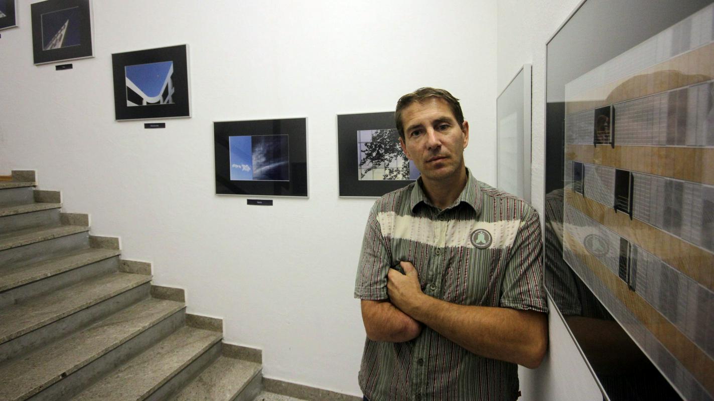 Klinični psiholog in fotograf Matej Peljhan s prekrižanimi rokami stoji ob razstavi fotografij. Na njegovi levi so kamnite stopnice, nad njimi se vrstijo različne umetniške fotografije. Foto: osebni arhiv