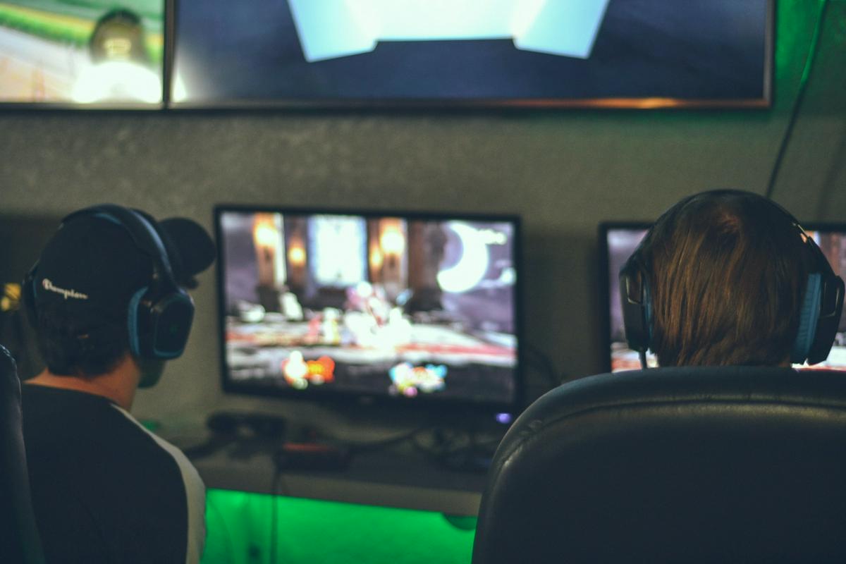 Dva igričarja s slušalkami sedita na stolu, obrnjena proti zaslonu, na katerem je zamegljena podoba živahne videoigre. Foto: Pixabay