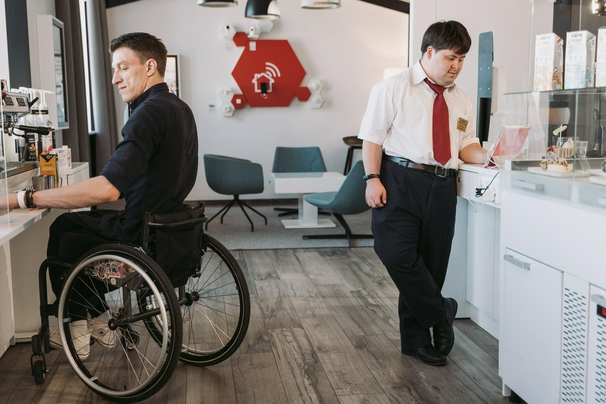 Dostopno urejena kavarna. Ob kavnem avtomatu moški na invalidskem vozičku, na nasprotni strani drug moški stoji ob pultu. Foto: pexels.com/Mart Productions