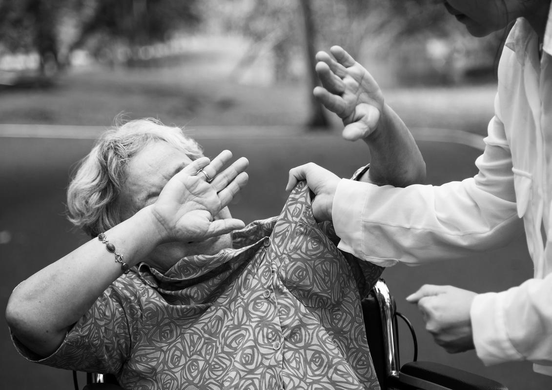 Na sliki je starejša ženska na invalidskem vozičku, ki drži roke pred obrazom, ker jo negovalka želi udariti. Foto: Shutterstock