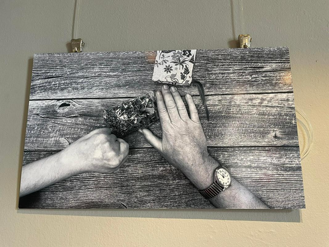 Na sliki je tloris rok položenih na leseno mizo. Fotografija je črno-bela. Leva roka je popolnoma stisnjena v pest, desna ima vse prste iztegnjene in uro na zapestju. Med rokama je prozorna vrečka s čajem. Foto: MMC RTV SLO/Ana Vojnović Zorman
