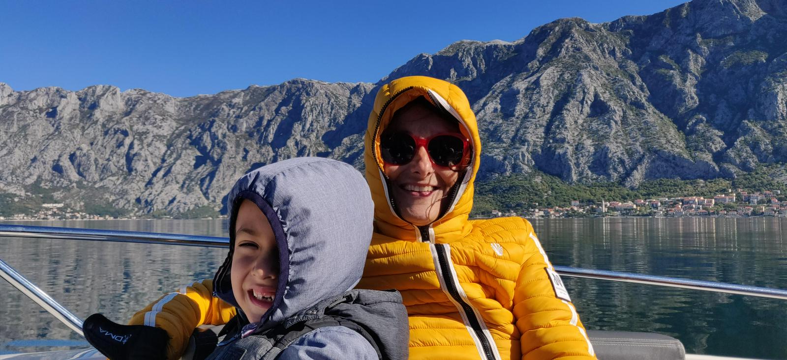 Na fotografiji sta Nina Wabra Jakič in Leo. Sta na čolnu sredi jezera, ki ga obdajajo gore. Oba nosita zimski jakni in sta si nadela kapuci, Nina nosi velika sončna očala z rdečim okvirjem. Oba sta nasmejana. Foto: Osebni arhiv Nine Wabra Jakič