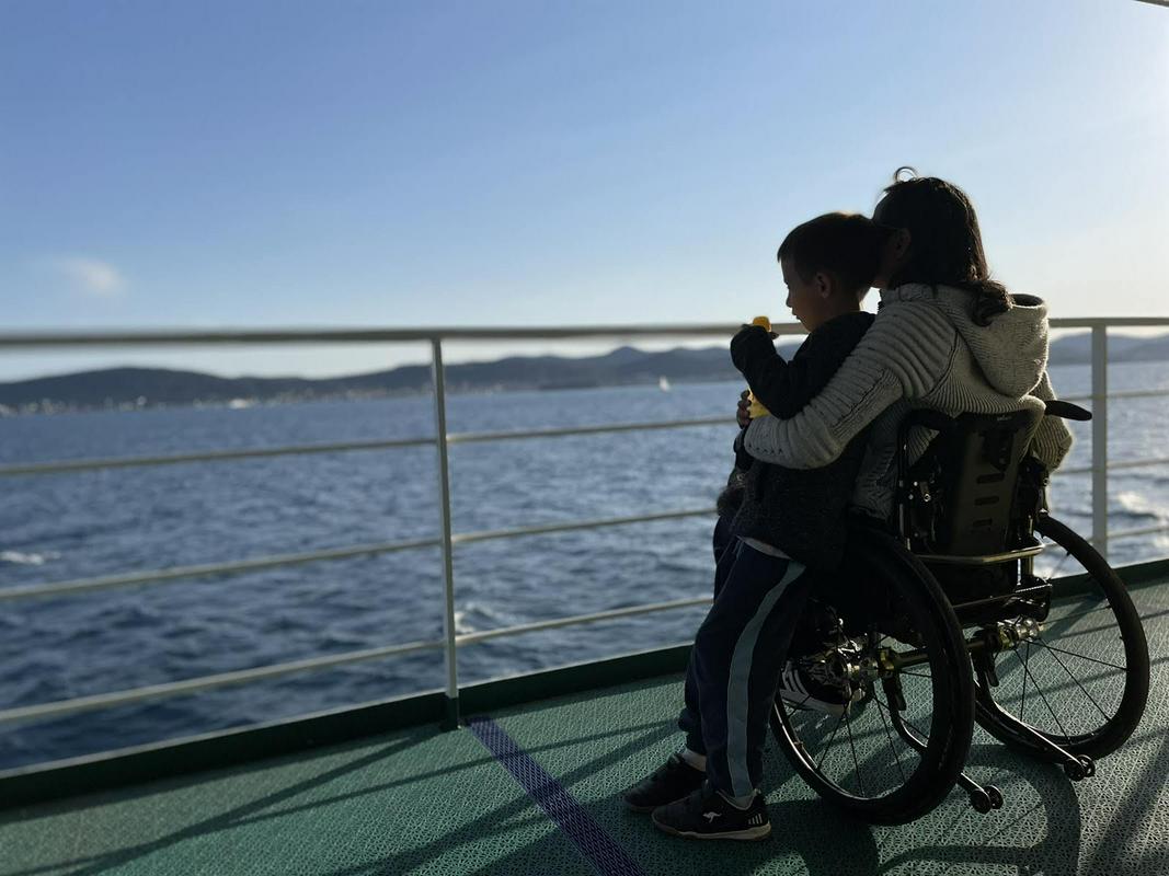 Na sliki sta Nina Wabra Jakič in Leo. Nina sedi na invalidskem vozičku in od zadaj objema Lea, ki stoji ob njej. Sta na trajektu in gledata morje in modro nebo. Foto: Osebni arhiv Nine Wabra Jakič