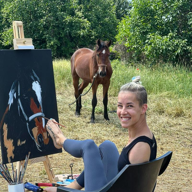 Na fotografiji je Dejana Bačko med slikanjem konja. Z levim stopalom, v katerem drži čopič, slika po platnu. Nahaja se nekje v naravi, pred njo pa pozira pravi konj. Foto: osebni arhiv Dejane Bačko