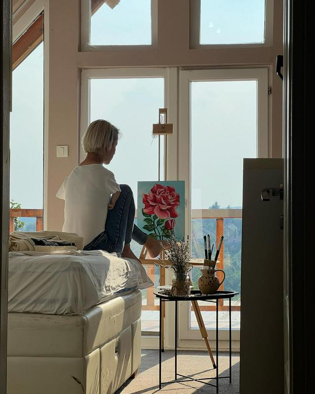 Dejana Bačko sedi na robu zakonske postelje in z levim stopalom slika motiv vrtnice. Ob njen je mizica, na kateri sta vaza s šopkom sivke in lonček s čopiči. V sobi je prijetna svetloba, ki prihaja skozi okno in steklena balkonska vrata pred njo. Foto: osebni arhiv Dejane Bačko