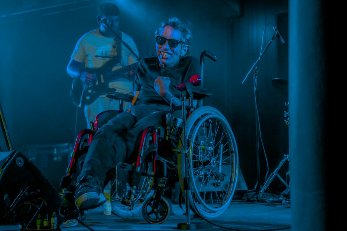 Na fotografiji je Fausto Sousa med koncertom v Kinu Šiška. Sedi na invalidskem vozičku in nosi sončna očala ter se prešerno smeji. Za njim stoji Jorge Maleiro in igra električno kitaro. Foto: Marcel Obal/arhiv Kino Šiška