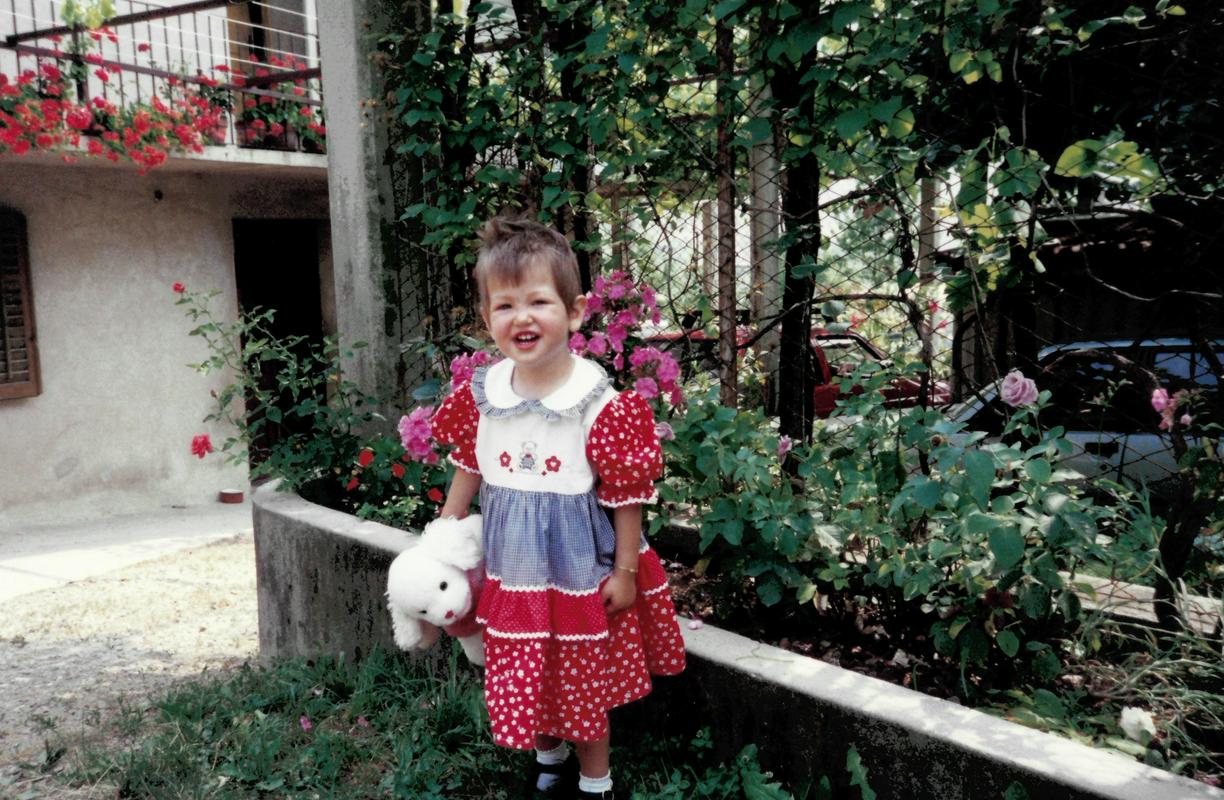 Vanessa stara 2 leti in pol na vrtu pred hišo. Stoji ob ograji, kjer rastejo vrtnice. Oblečeno ima rdečo oblekico z belimi pikicmi in ovratnikom. V roki drži belega puhastega zajčka. Ima kratke rjave lase in se smeje. Foto: Osebni arhiv