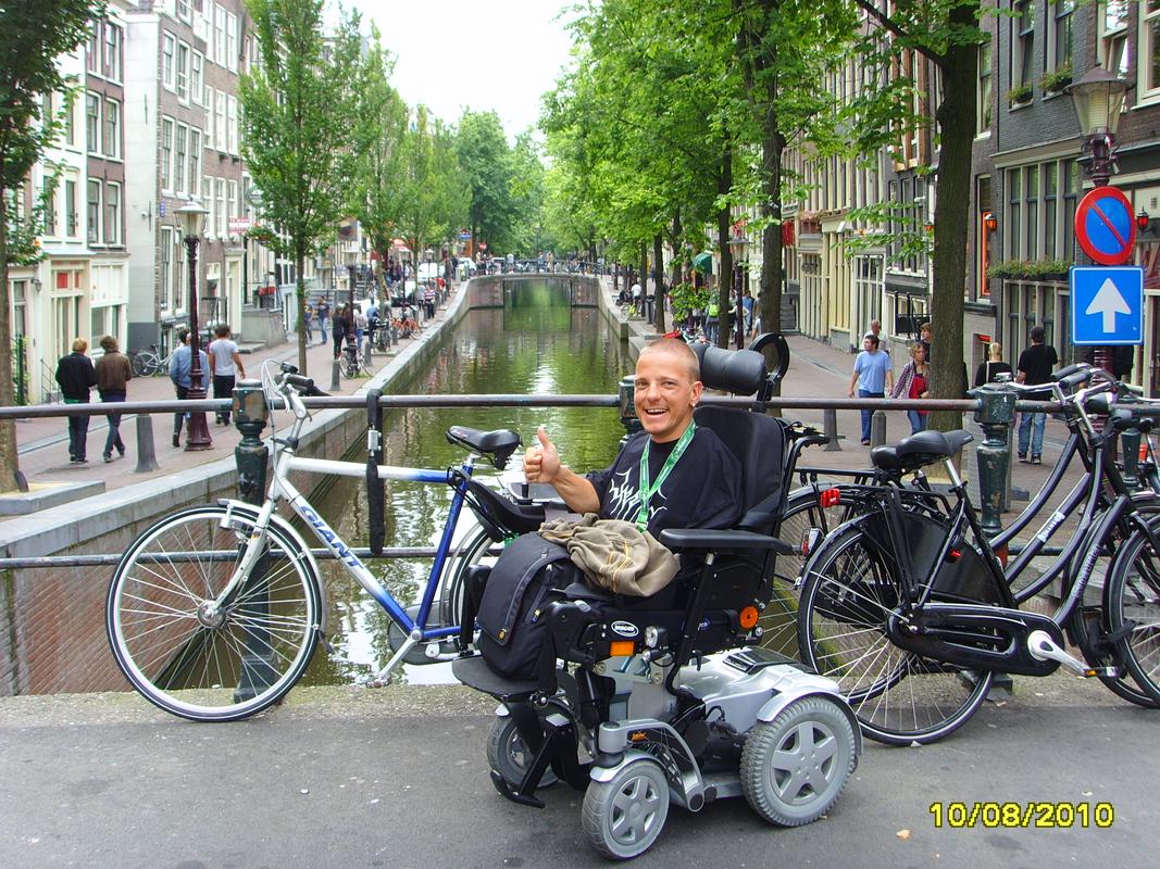 Miha Nardoni v Amsterdamu na enem od številnih mostičkov čez reko Amstel. Sedi na električnem vozičku in se z dvignjenim palcem smeji. Okoli njega so parkirana kolesa, v ozadju je sprehajališče, ki poteka na vsaki strani reke, ki jo obdaja in drevored. Foto: Osebni arhiv