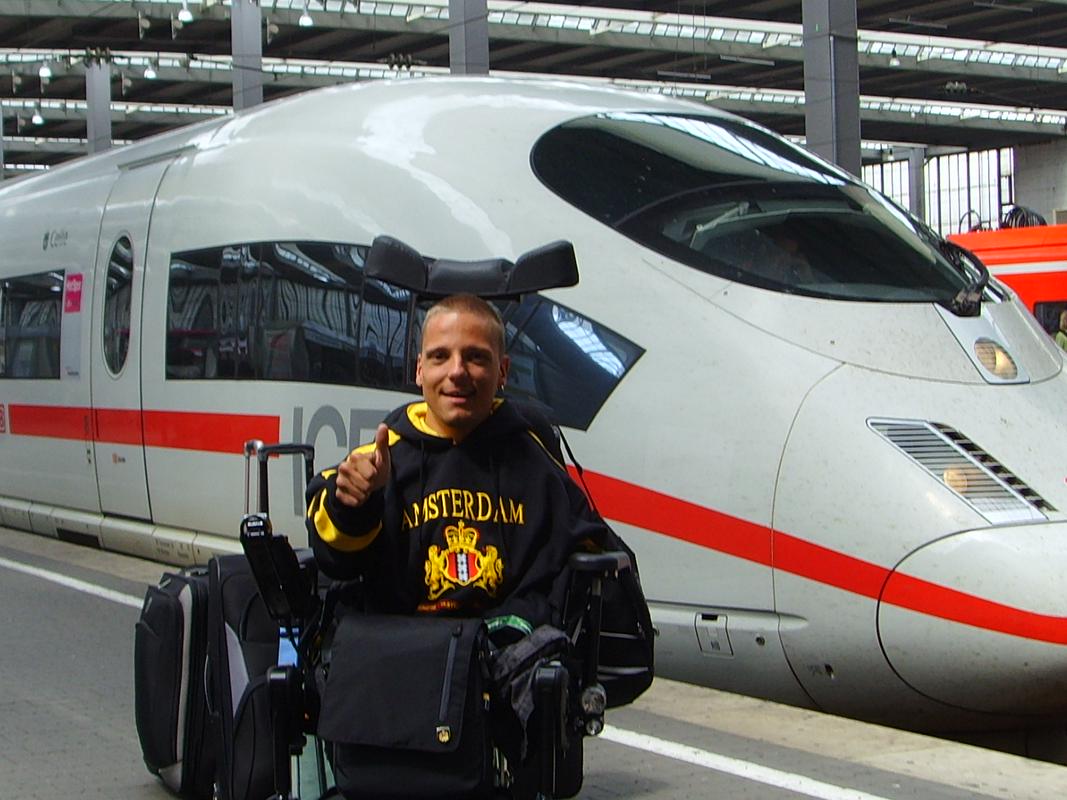 Miha na železniški postaji na poti v Amsterdam pred 14 leti. V ozadju je hitri vlak bele barve z rdečo črto. Miha dviguje palec na roku in se smehlja. Foto: Osebni arhiv